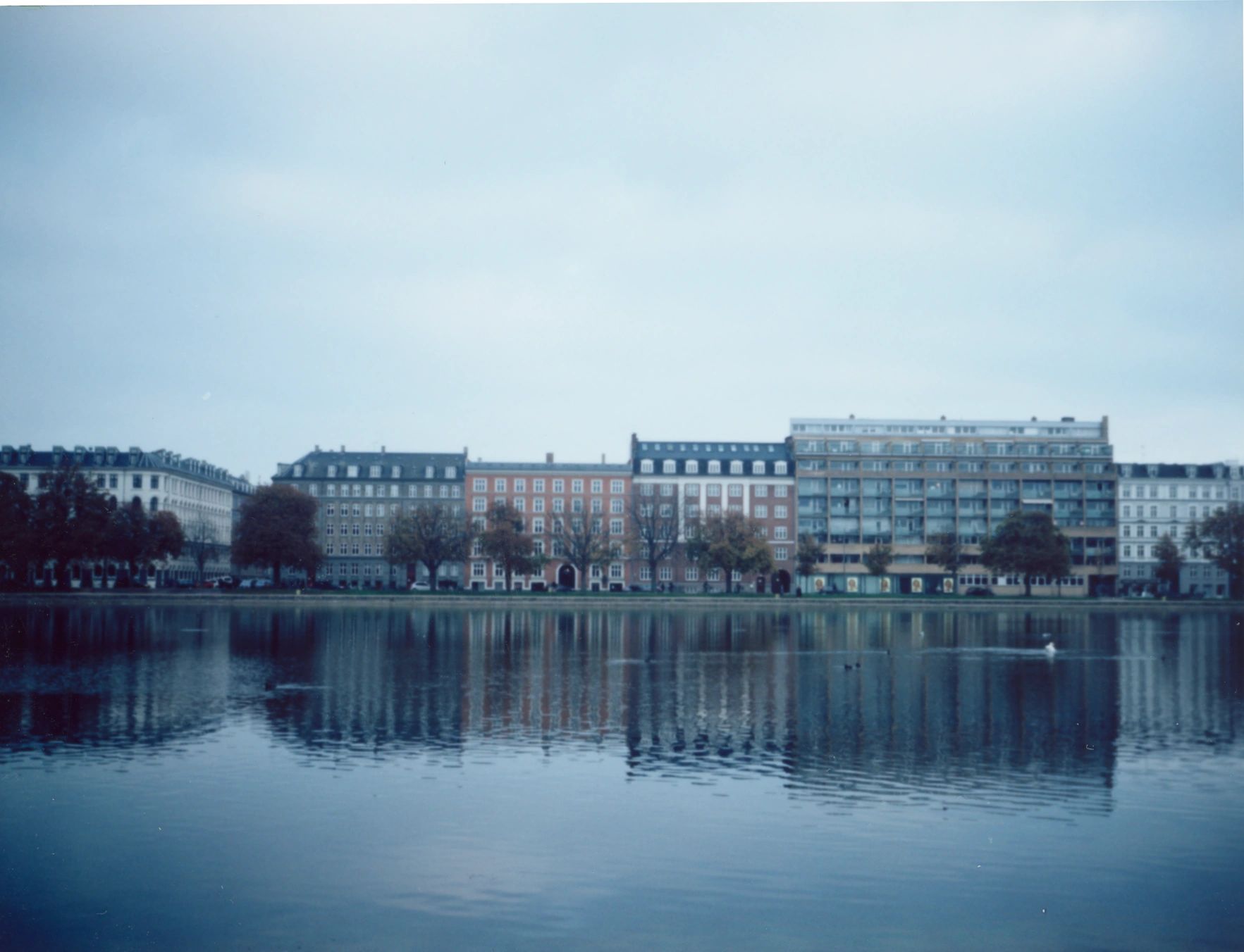Calm waterfront view reflecting a row of colorful buildings under an overcast sky.