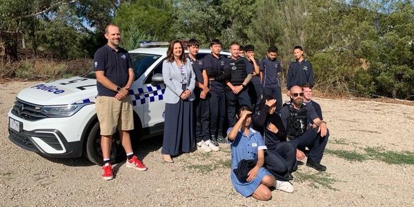 Group of police officers and students posing outdoors with a police vehicle.