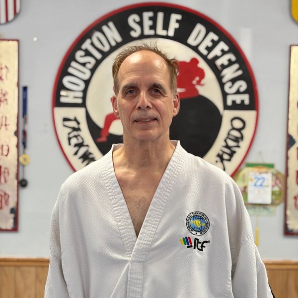 Man in a martial arts uniform standing in front of a Houston Self Defense sign.