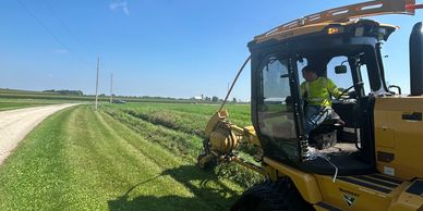 A man operates a yellow mower machine cutting grass beside a rural road on a sunny day.