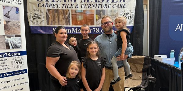 Family team at Steam Master cleaning service booth with promotional materials and candy bowl.