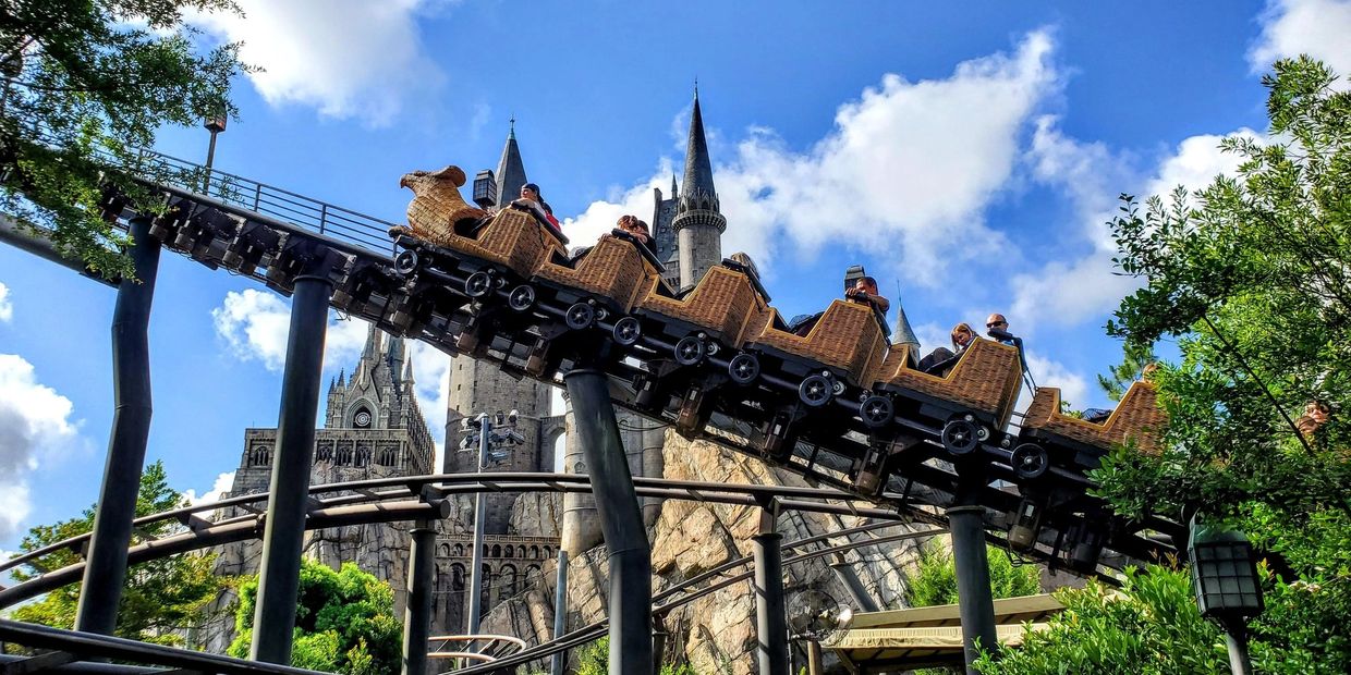 A roller coaster with wicker basket cars rides past a castle under a blue sky.