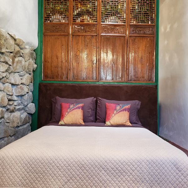 Cozy bed with decorative pillows against a rustic wooden panel and stone wall.