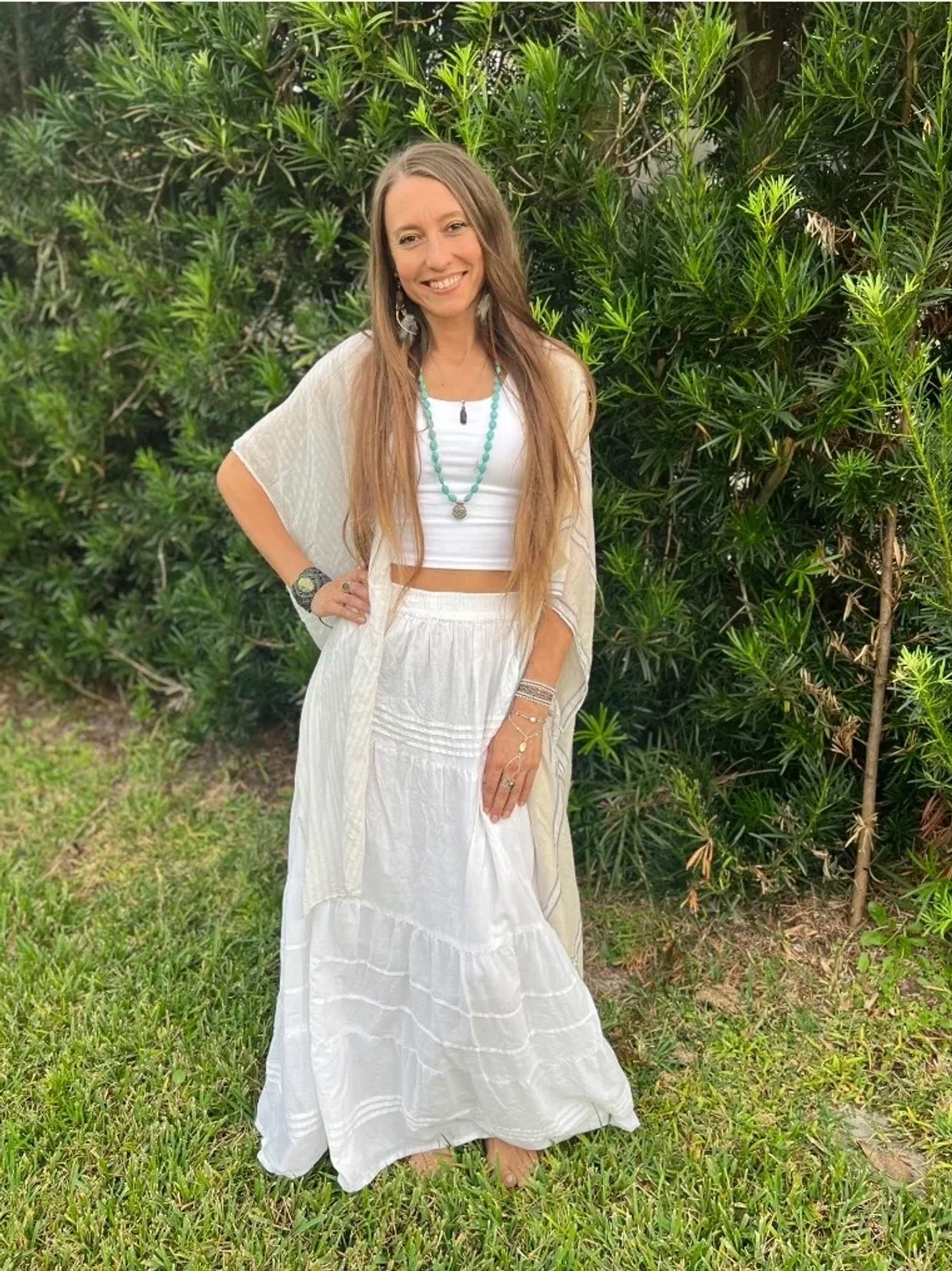 A woman in a white outfit smiles outdoors against lush greenery.