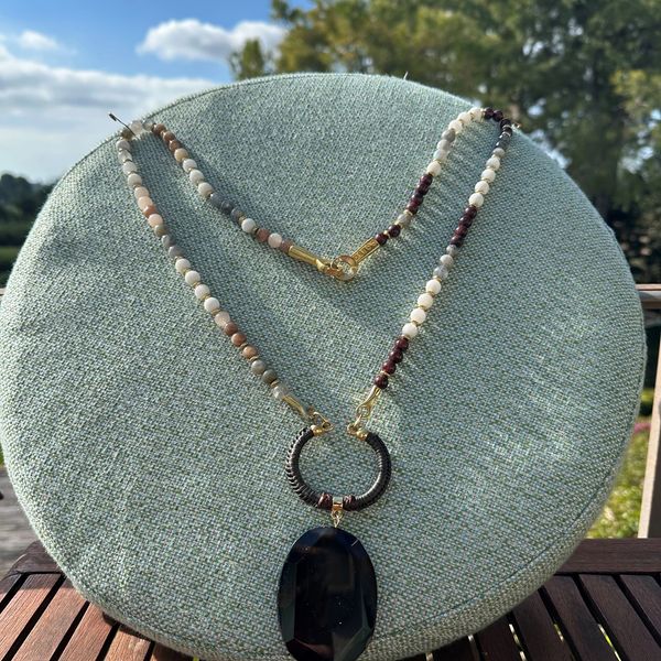 Beaded necklace with a large black pendant displayed outdoors on a green stand.