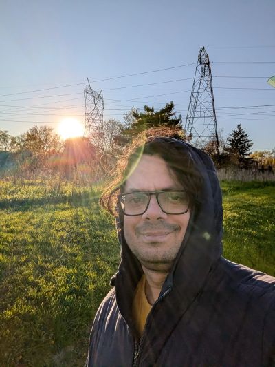 Man in a hooded jacket smiling outdoors at sunset with power lines in the background.