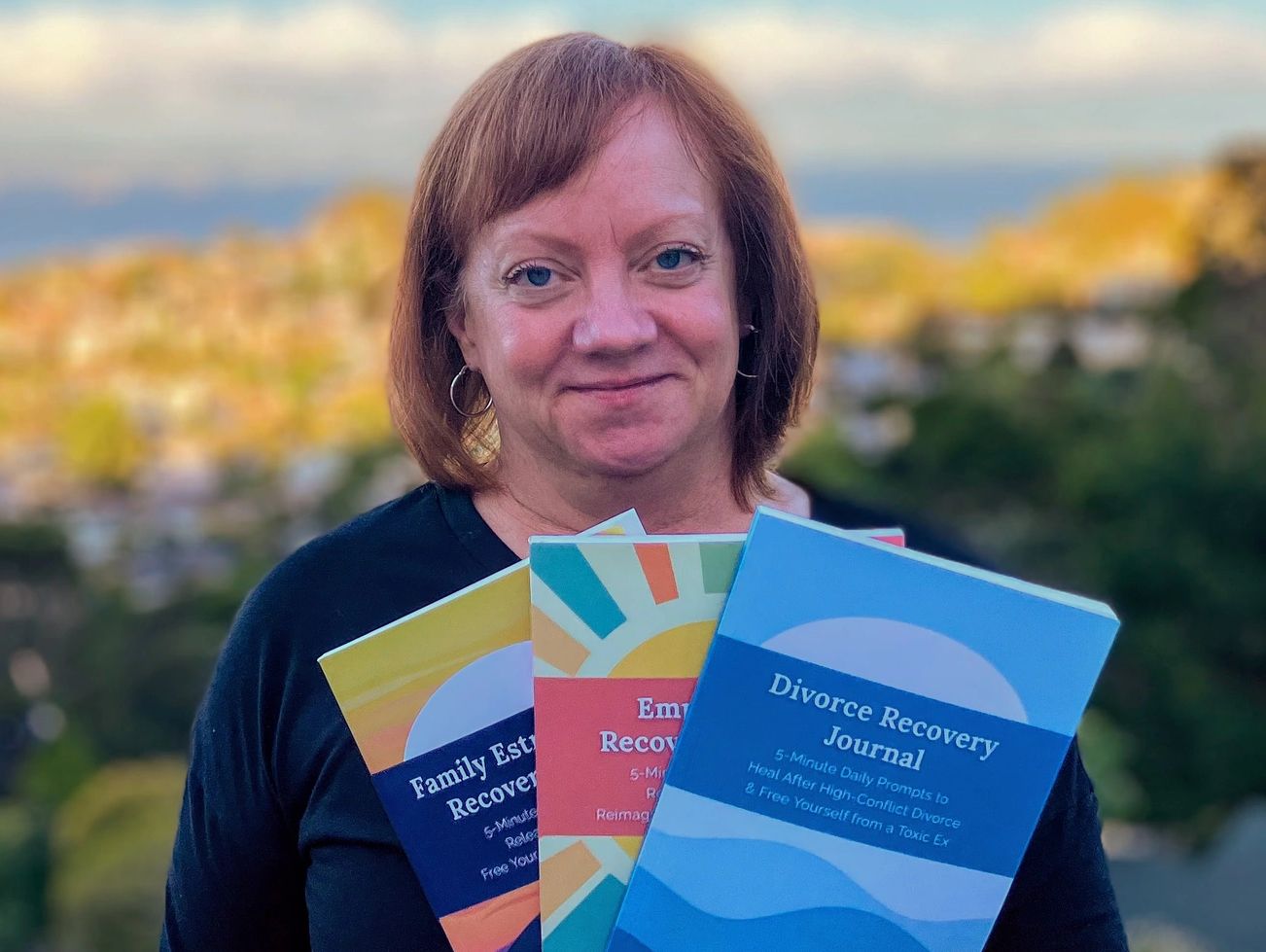 Woman holding three colorful recovery journals outdoors with a smile.