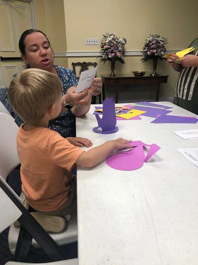 A woman helps a child with a colorful paper craft at a table.