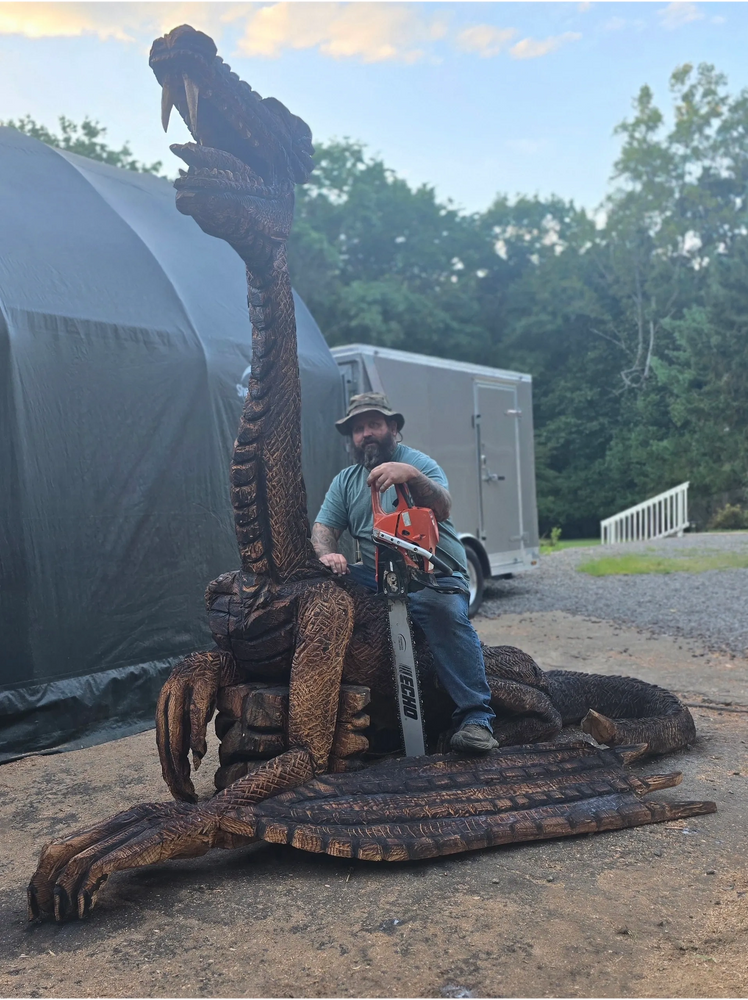 Man with chainsaw sitting on a large wooden dragon sculpture outdoors.