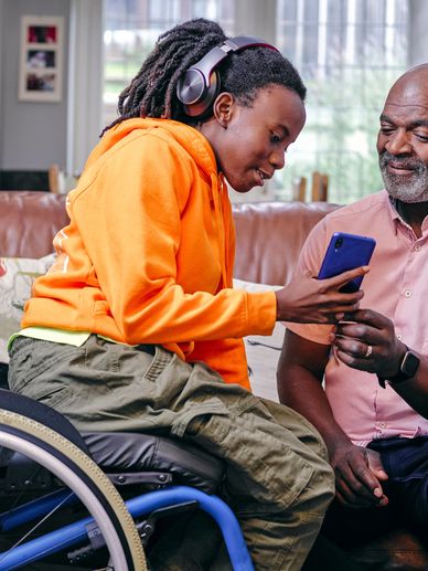 Young person in wheelchair showing something on phone to older man on couch.