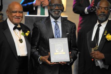 Three men in tuxedos at an awards ceremony, one holding a plaque.