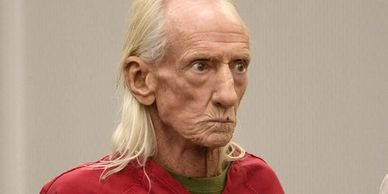 Elderly man with long white hair wearing a red jacket.