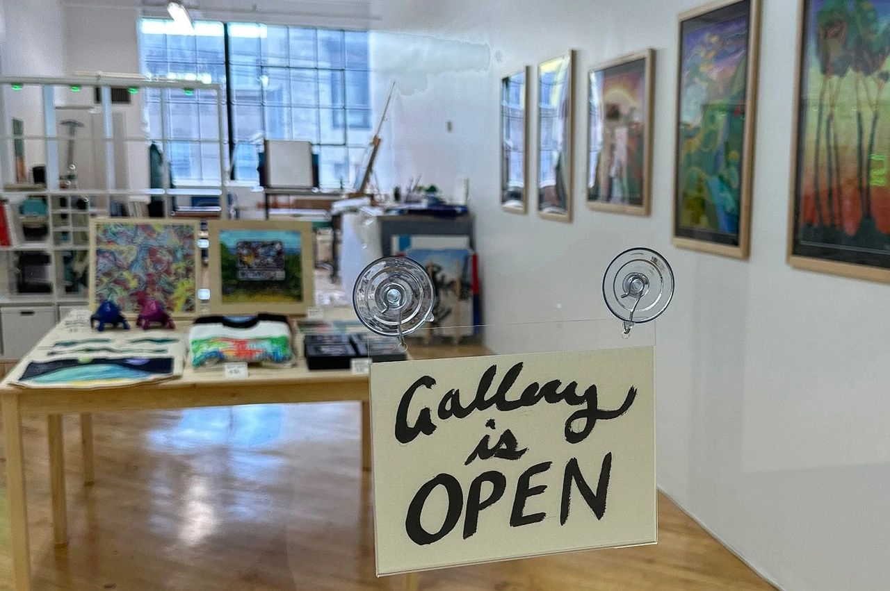A sign reading 'Gallery is OPEN' in front of a gallery space featuring colorful artwork and various art supplies displayed on a wooden table.