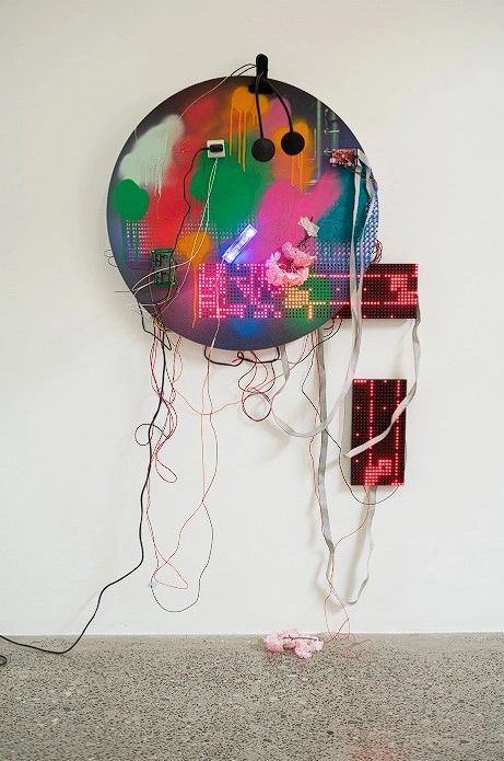 An abstract wall installation featuring a round, colorful piece with spray paint patterns and electronic components, including visible wires and LED display panels, mounted against a plain wall.