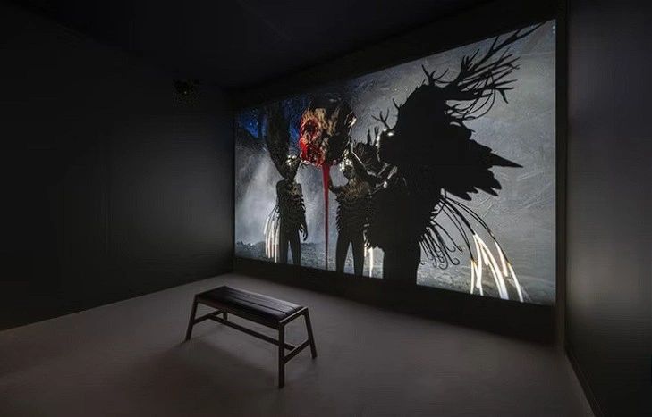 A darkened gallery featuring a large projection on the wall depicting shadowy figures with exaggerated wings and a central figure with a dramatic, colorful design. A wooden bench is positioned in front of the display.