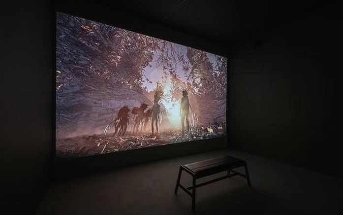 An art installation featuring a projection on a large screen depicting shadowy figures in a forest scene, with a bench in the foreground for viewers.