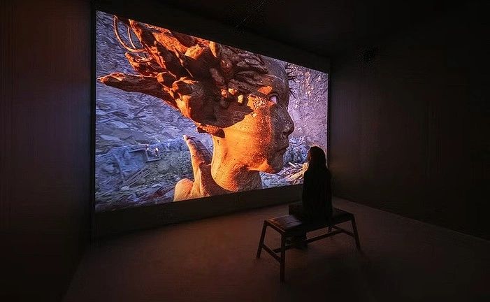 A person sitting on a bench in a dark room, watching a large screen displaying an artistic video featuring a stylized human face and abstract, natural elements.