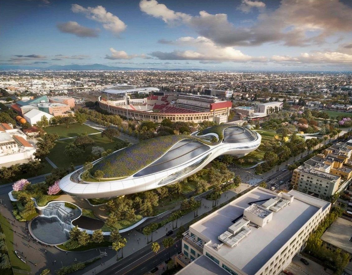 Aerial view of a futuristic building with unique, flowing architecture surrounded by greenery, next to a sports stadium and urban landscape.