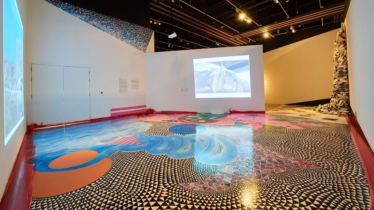 An art installation featuring an intricately designed floor with colorful patterns, a video projection on the wall, and sculptural elements in a contemporary gallery setting.