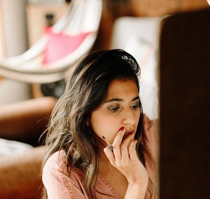 A woman with long dark hair is seated thoughtfully, resting her chin on her hand while looking down. She has a cigarette in her hand and is in a cozy indoor setting.