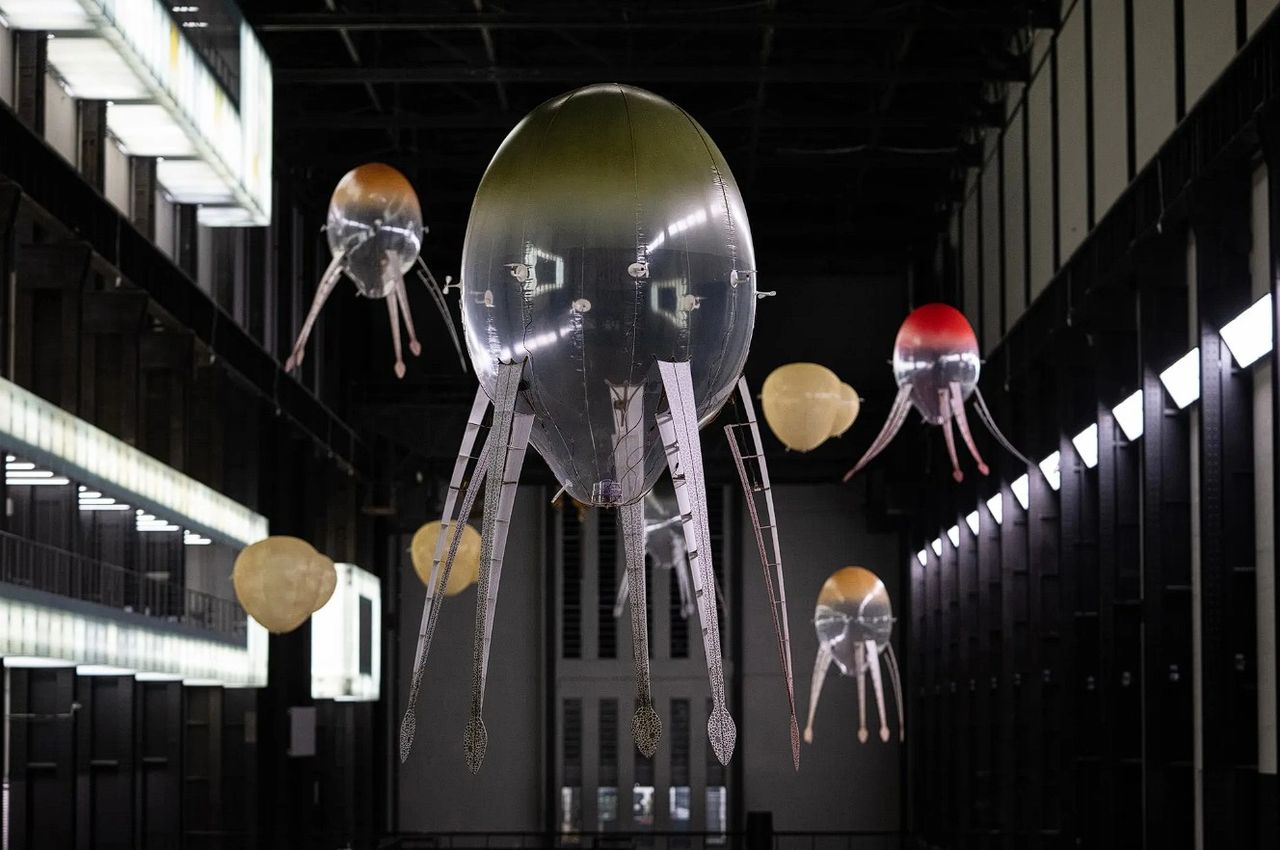 An art installation featuring large, floating, transparent jellyfish-like sculptures with colorful accents, suspended in a spacious industrial setting.