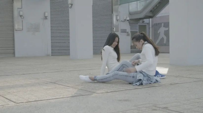 Two young women sitting on the ground outdoors, engaged in conversation or interaction, with one adjusting her clothing. They are in a modern urban setting with a minimalist background.