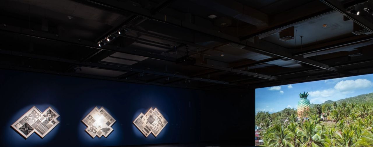 An art exhibition space featuring three illuminated wall pieces against a dark blue wall, alongside a large projection of a tropical landscape with a pineapple structure among palm trees.