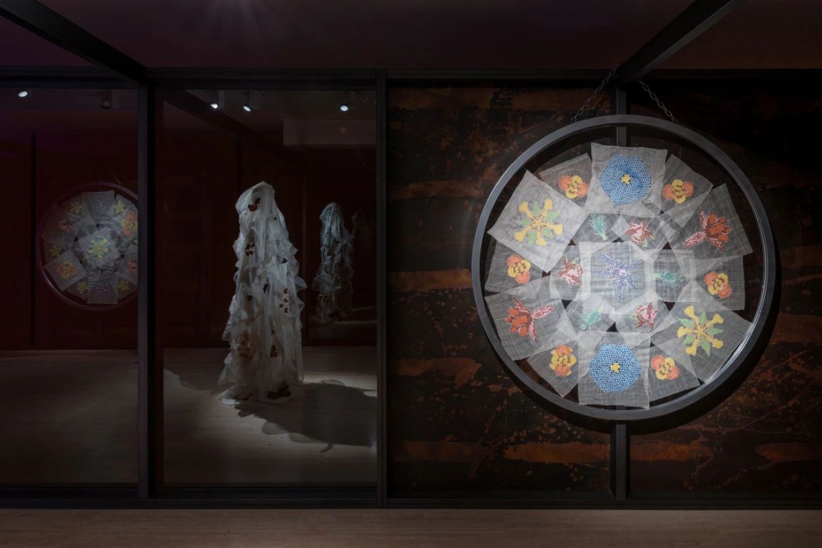 An art installation featuring a large circular embroidery display and a ghostly white figure draped in fabric, set against a dark background in a gallery space.