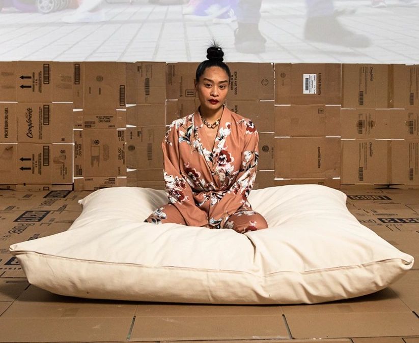A person sitting cross-legged on a large cushion, wearing a floral robe, with a backdrop of stacked cardboard boxes.