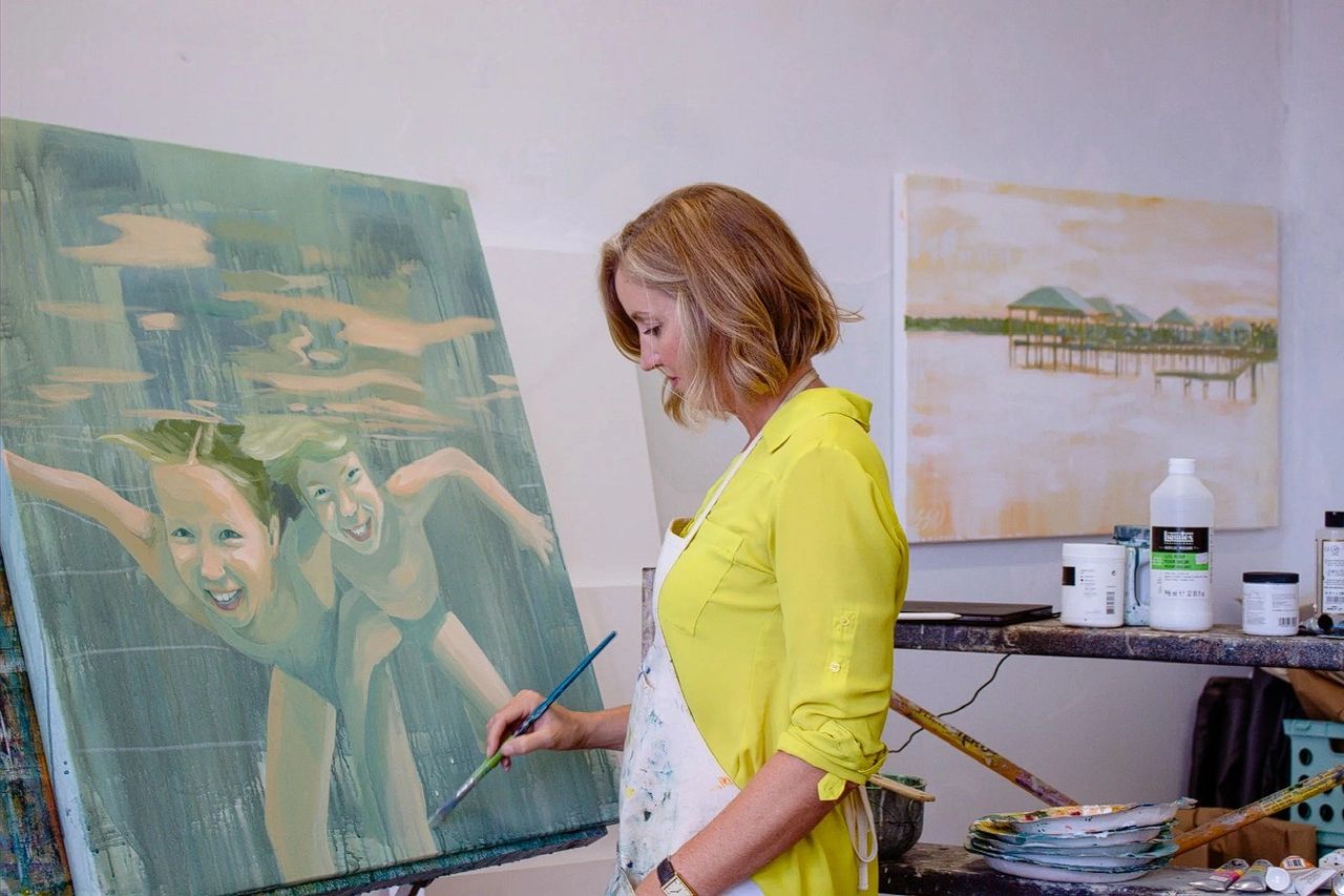 An artist in a yellow shirt painting a large canvas featuring two children swimming underwater in an art studio.