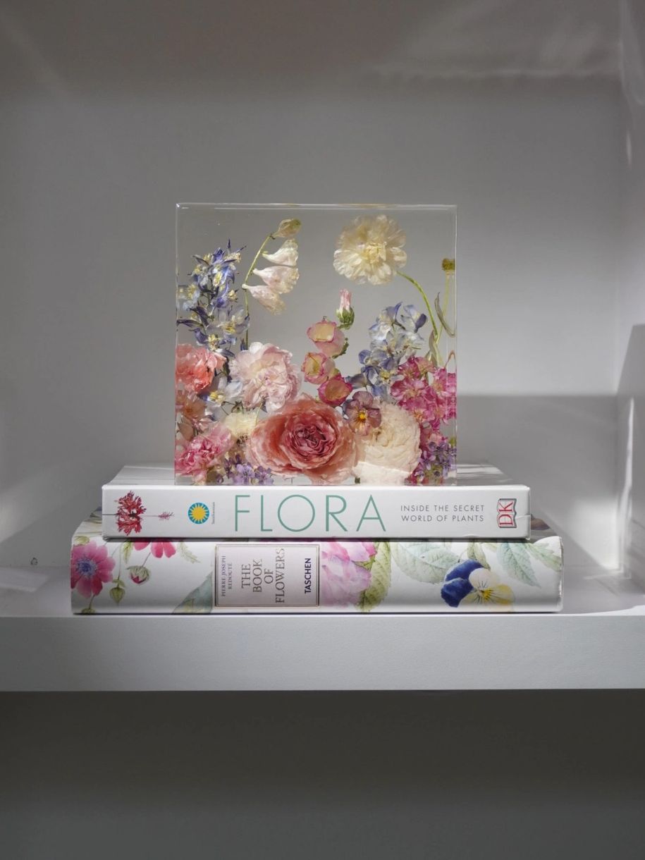 Decorative block with pressed flowers atop floral-themed books on a white shelf.
