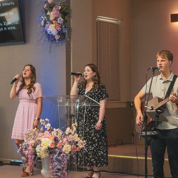 Three young people singing and playing guitar on stage with flowers.