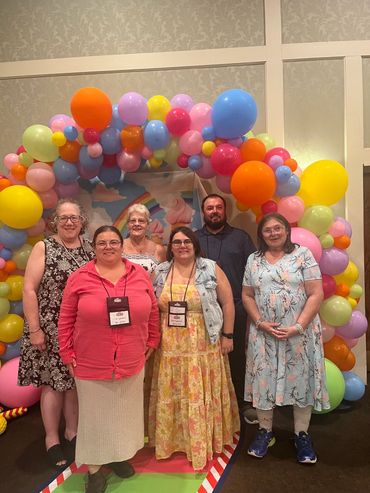 Six people posing in front of a colorful balloon arch.