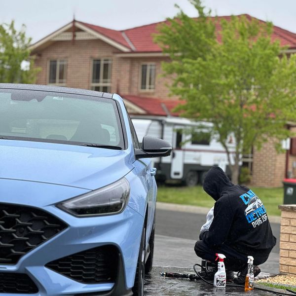 Person cleaning a blue car in a residential driveway on a rainy day.
