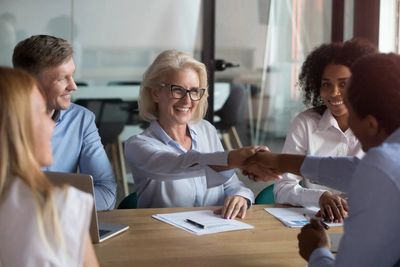 Five people dressed in business attire around a table with papers & laptop. Two are shaking hands.