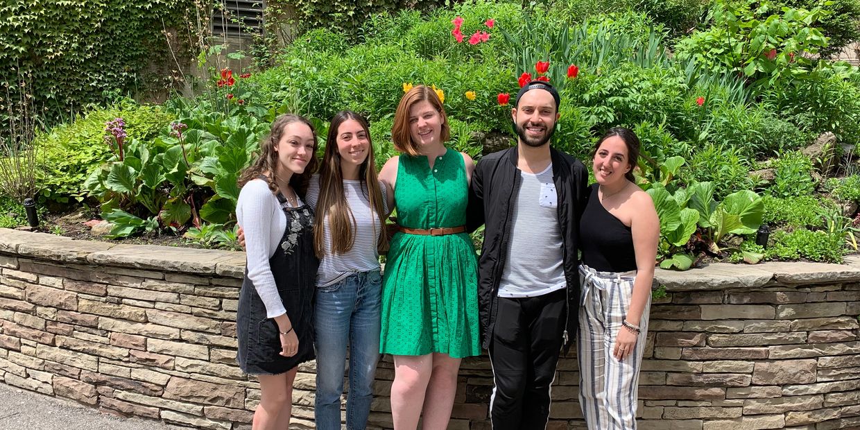 Taylor Medeiros, Anna Zappone, Emma Graham, Adam Gomes, and Jessica Sorbara pose together in front of a bed of flowers outside Brennan Hall. 