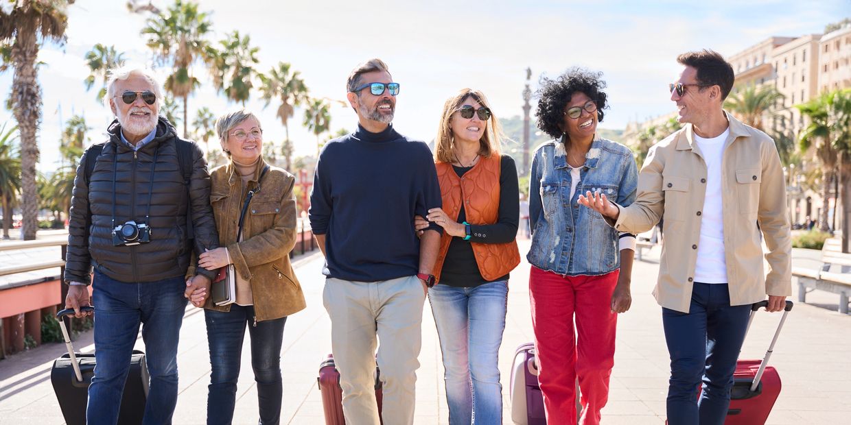 Six friends walk down a sidewalk together, each pulling a suitcase behind them. 