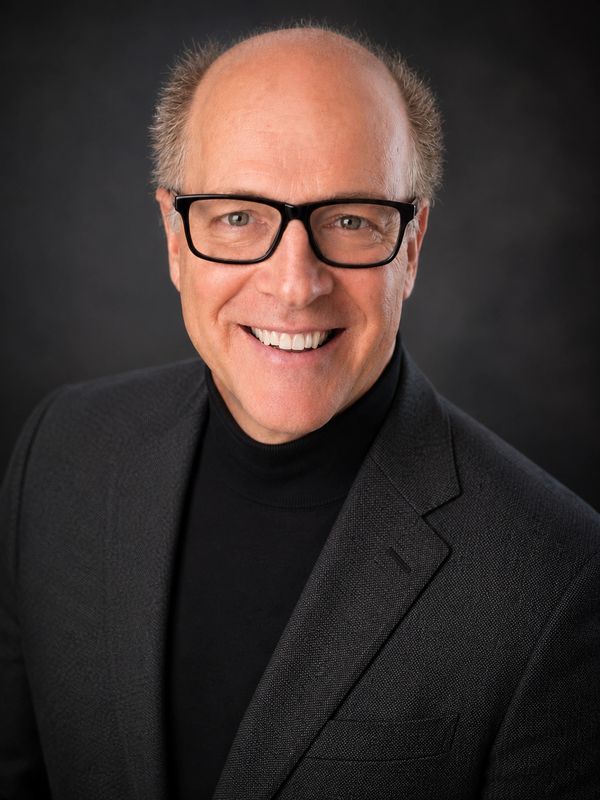 Smiling bald man with glasses in a dark suit jacket and black turtleneck against a dark background.