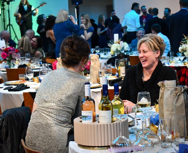 2 women are seated at a table talking and laughing. There is a large crowd behind them.