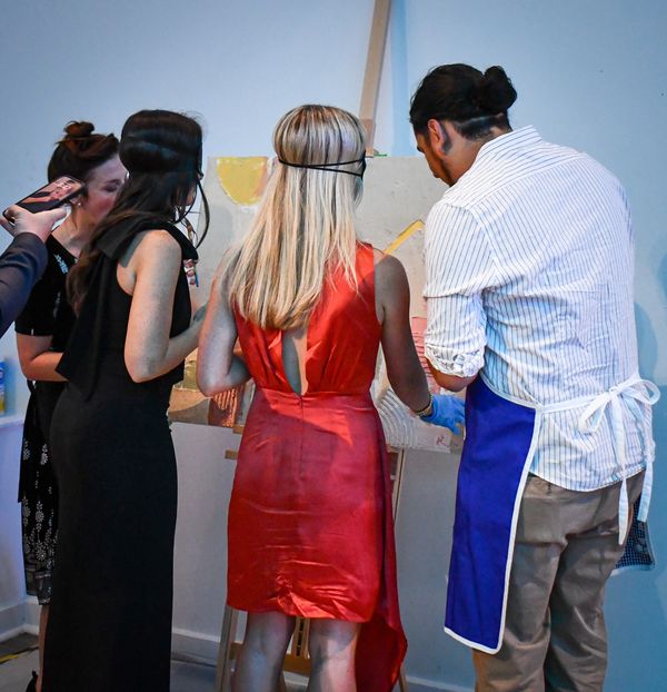 4 people standing in front of a canvas. A blond woman in a red dress is painting while blindfolded.