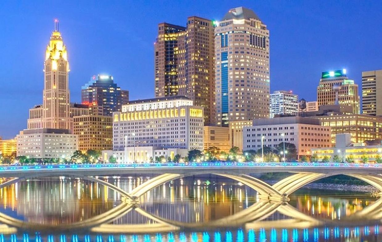 City skyline at dusk with illuminated buildings and a lighted bridge reflecting on water.
