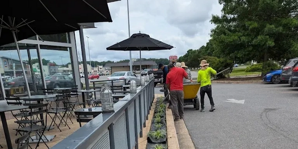 Workers planting greenery along a sidewalk near outdoor seating and parking lot.