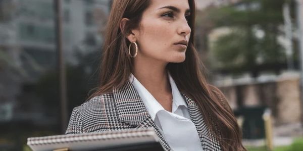 Woman in a checkered blazer looking thoughtfully to the side outdoors.