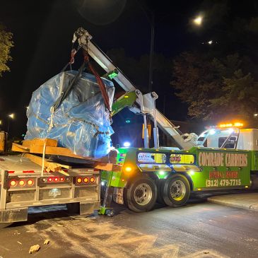 Tow truck lifting a large wrapped object onto a flatbed at night.