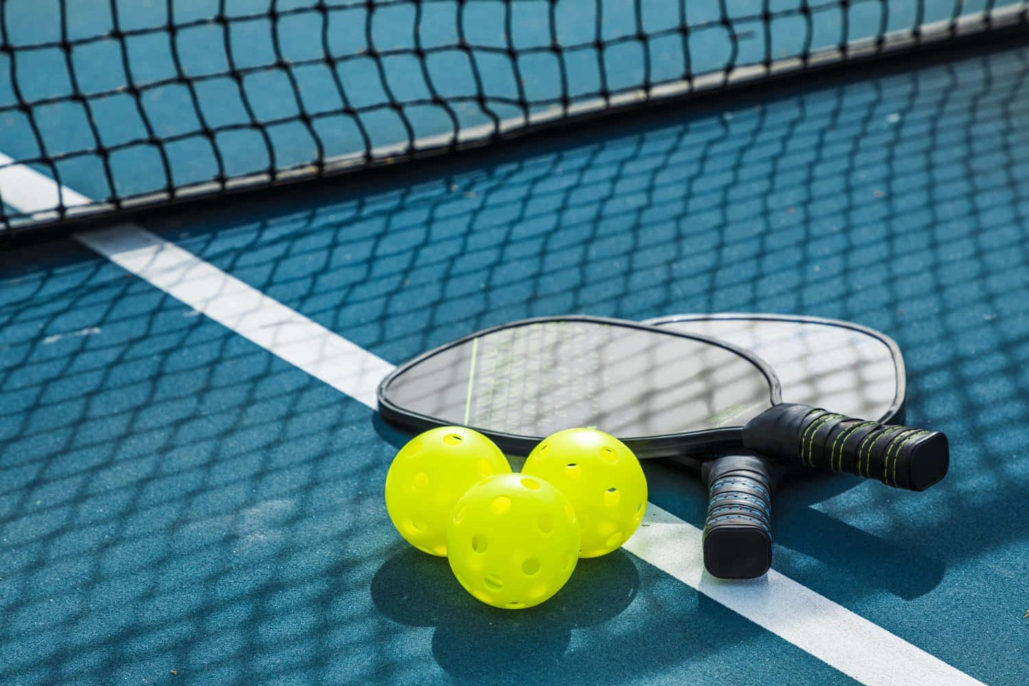 Two pickleball paddles and three yellow pickleballs on a blue court near the net.