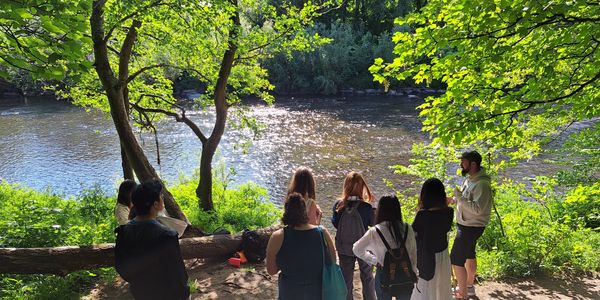 Group of people gathered by a sunlit river surrounded by lush greenery.
