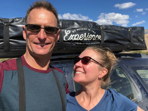 A smiling couple wearing sunglasses stands in front of a vehicle on a sunny day.