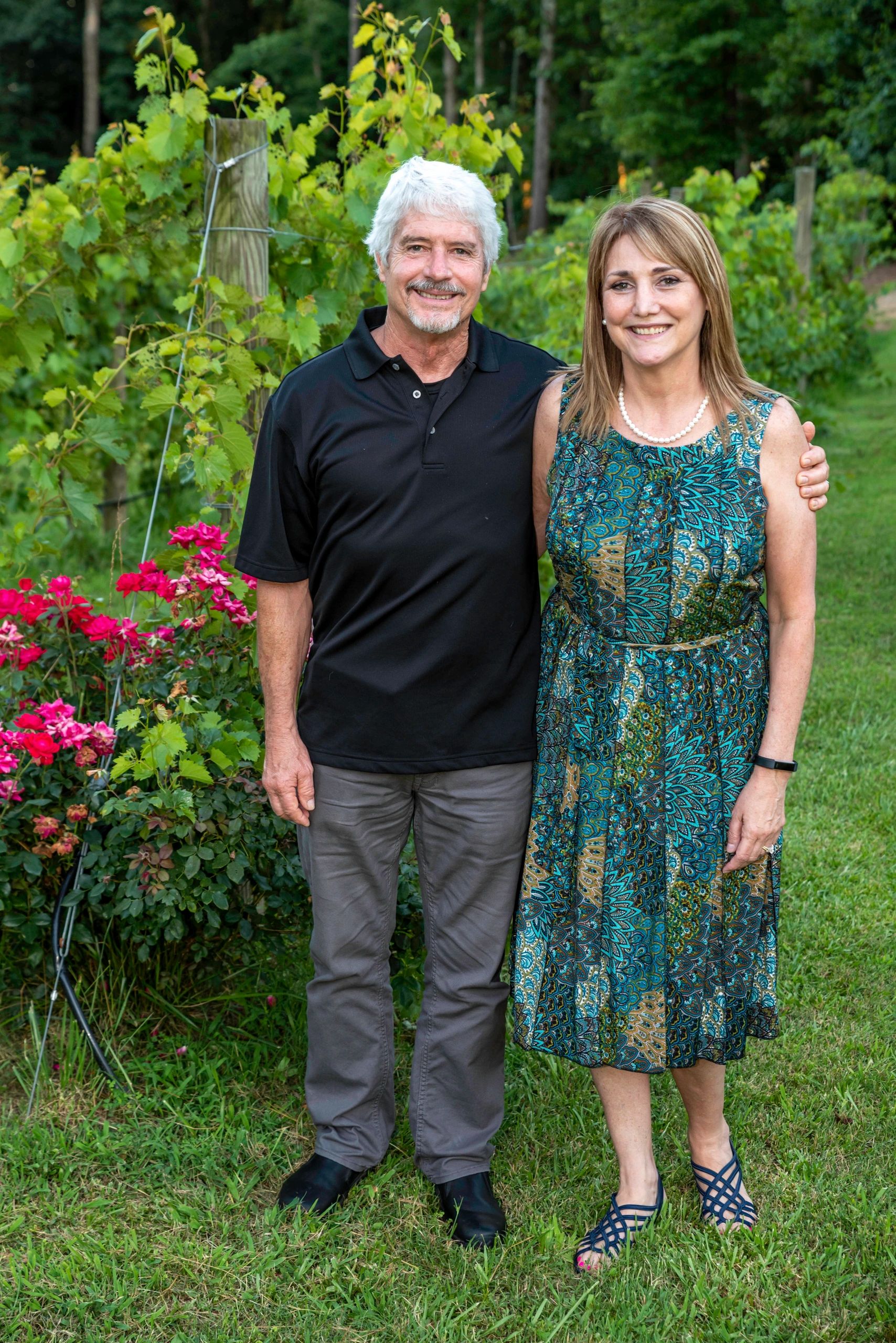 A smiling couple stands in a garden with green plants and pink flowers.