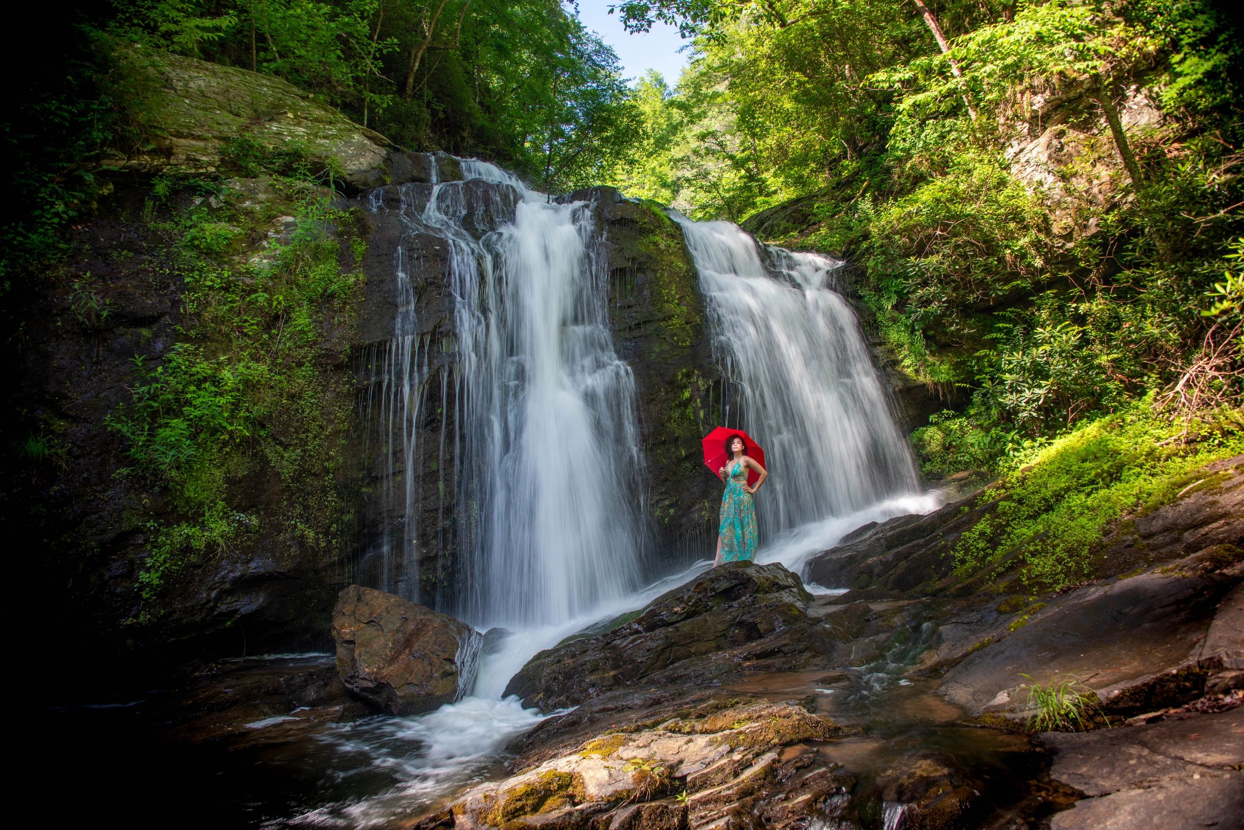 A woman with a red umbrella stands near a waterfall in a lush forest.