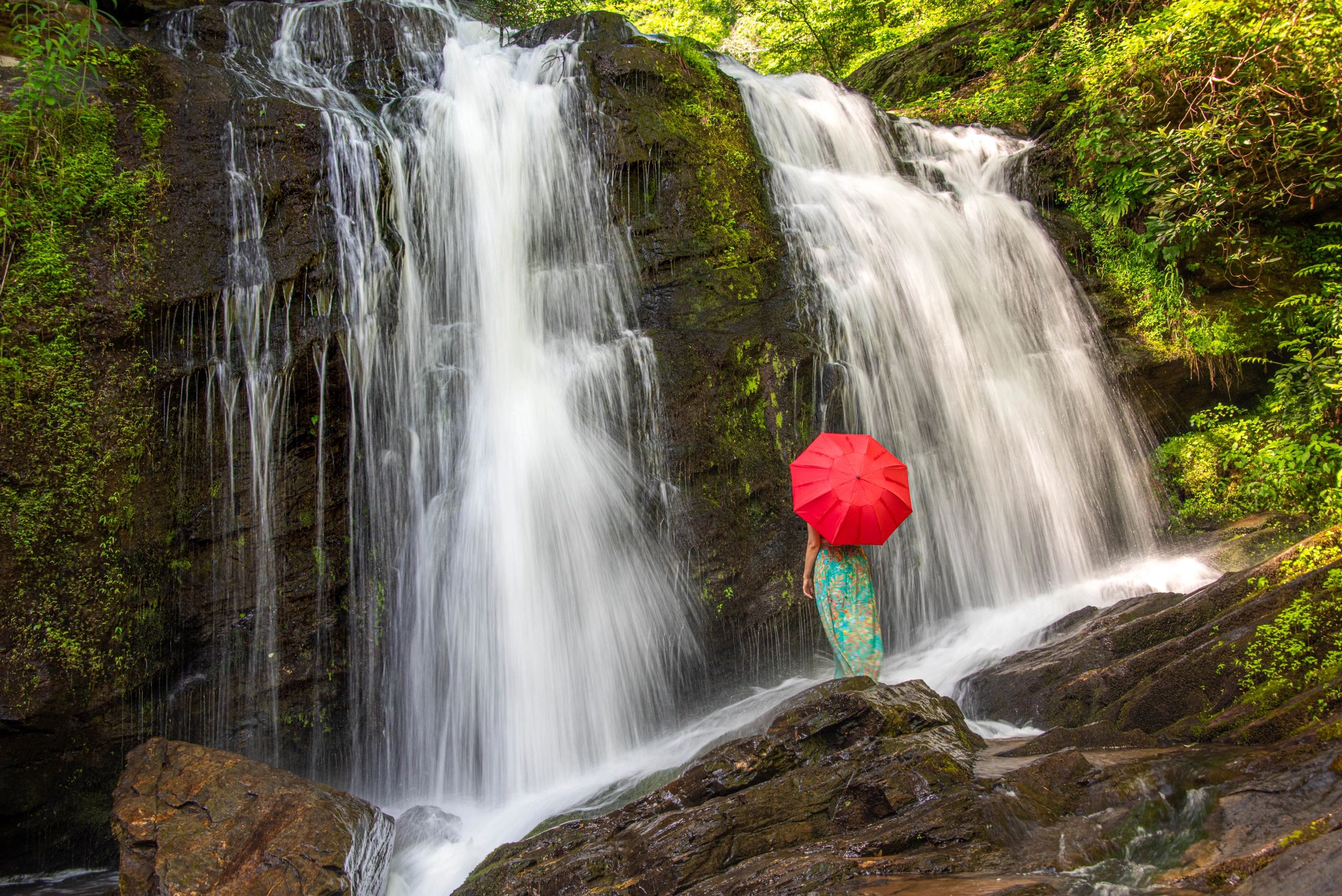 Person with red umbrella standing near a lush waterfall.
